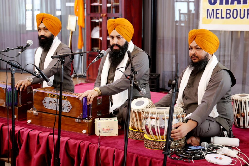 Three Sikh musicians playing harmoniums and tabla performing kirtan indoors.