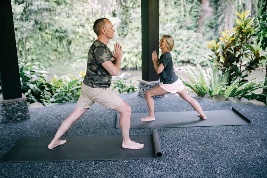 Couple practicing yoga outdoors on mats surrounded by lush greenery.