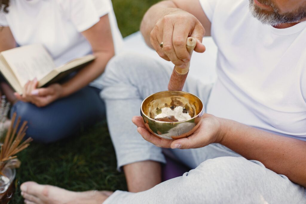 A man meditates outdoors using a Tibetan singing bowl, seated with a book nearby.