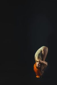 A redheaded woman performs a contortion pose against a black background in a studio setting.