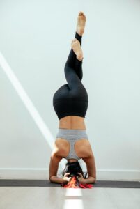 Full body of back view of anonymous barefoot female in sportwear performing supported headstand Garudasana on mat against white wall