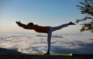 Person practicing yoga in warrior pose above a scenic cloudscape during sunrise.