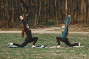 Two women in masks doing yoga poses outdoors amidst COVID-19 precautions.
