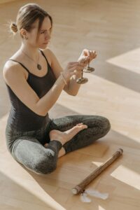 Adult woman meditating on wooden floor holding tingsha cymbals in sunlight.